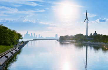 A tall wind turbine stands out across the river from a hazy city skyline. 